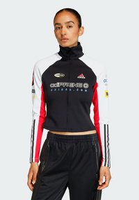 Crna i bijela skraćena jakna s crvenim naglascima, koja ima visoki ovratnik i zatvaranje na patentni zatvarač. "adiPRENE" oznaka prikazana je istaknuto.