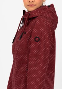 Bordeaux jack met rode stippen, voorzien van een capuchon, trekkoord en een zwarte cirkelvormige accent op de mouw. Zachte textuur en casual ontwerp.