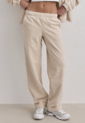 Persona vestida con pantalones deportivos beige de forro polar y zapatillas deportivas blancas, de pie con las manos en los bolsillos contra una pared lisa.