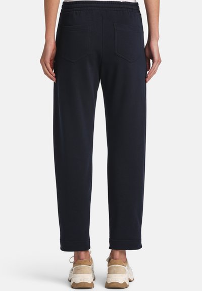 Person wearing navy blue relaxed-fit pants with elastic waistband and two back pockets, paired with beige and white sneakers, standing still.