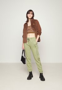 Brun mockakjacka med dragkedja, gröna sweatpants med dragsko och svarta ankelskor. Croppad topp med horisontella ränder i jordnära färger.