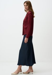 Blusa in raso bordeaux con maniche lunghe, abbinata a una lunga gonna blu navy. Il modello indossa décolleté beige con tacco, in piedi su uno sfondo neutro.