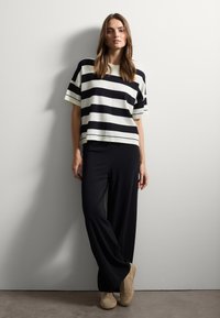 Une femme s'appuie contre un mur blanc, portant un haut à rayures horizontales noir et blanc à manches courtes, un pantalon noir à jambes larges et des baskets beiges.