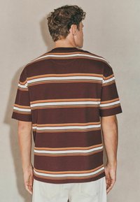 Kortærmet, maroon strikket skjorte med vandrette striber i orange og hvid. Har en afslappet pasform og en rund hals design.