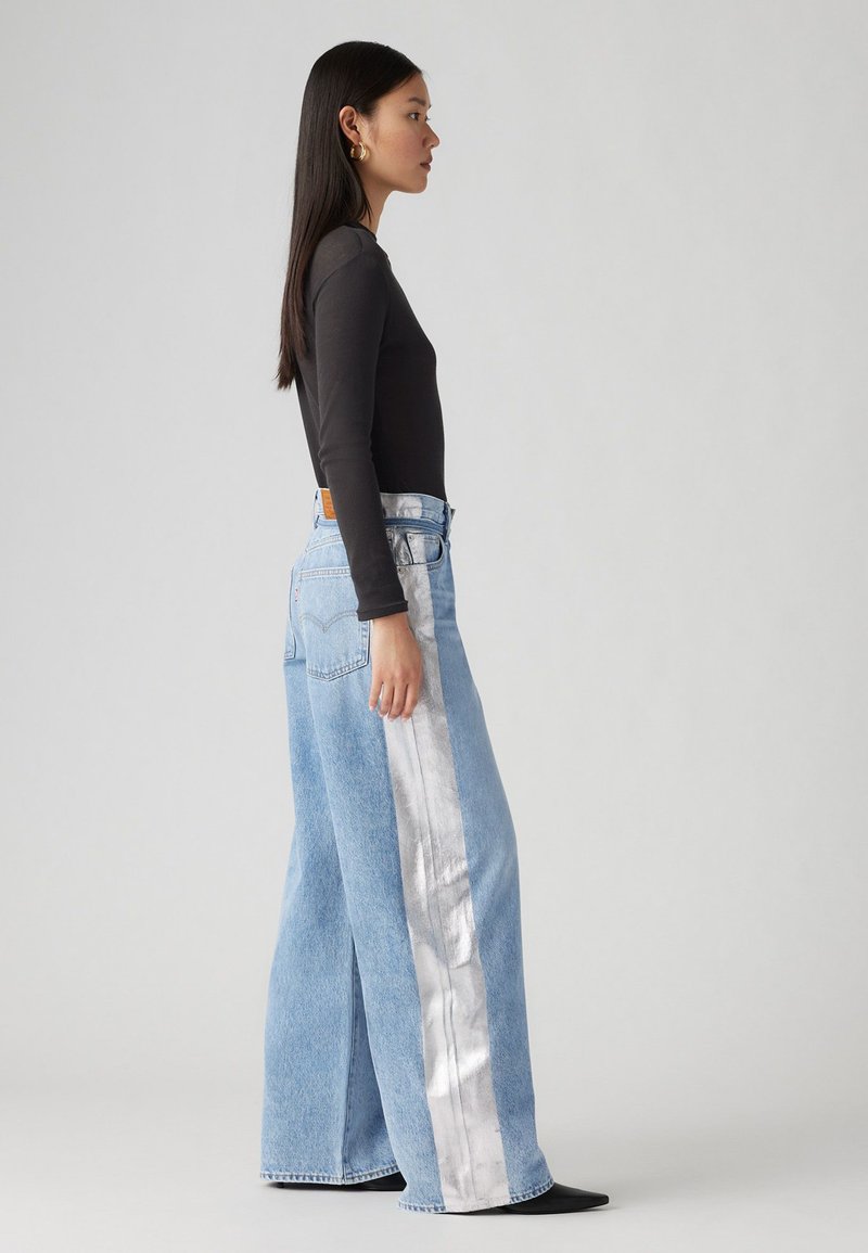 Jeans a gamba larga blu chiaro con una striscia laterale argentata, realizzati in denim, con vita alta e cinque tasche. Il modello indossa un top nero a maniche lunghe.