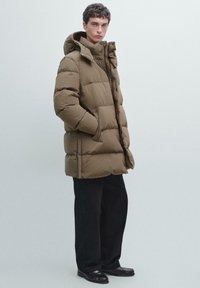 Giacca imbottita in verde oliva con design trapuntato, chiusura frontale con zip e cappuccio attaccato. Il materiale sembra leggero ma voluminoso.