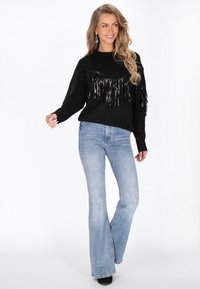 Maglione nero con frange e dettagli in paillettes sulla parte frontale, abbinato a jeans azzurri a zampa. Materiali e texture ad alto contrasto.