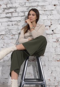 Chemise beige à manches longues et à côtes, pantalon ample vert foncé et bottes à lacets crème. Assise sur un tabouret en métal contre un mur en briques blanches.