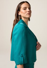 Blazer croppato turchese con colletto a rever, maniche a tre quarti, dettagli in bottone sulle maniche e una texture liscia.
