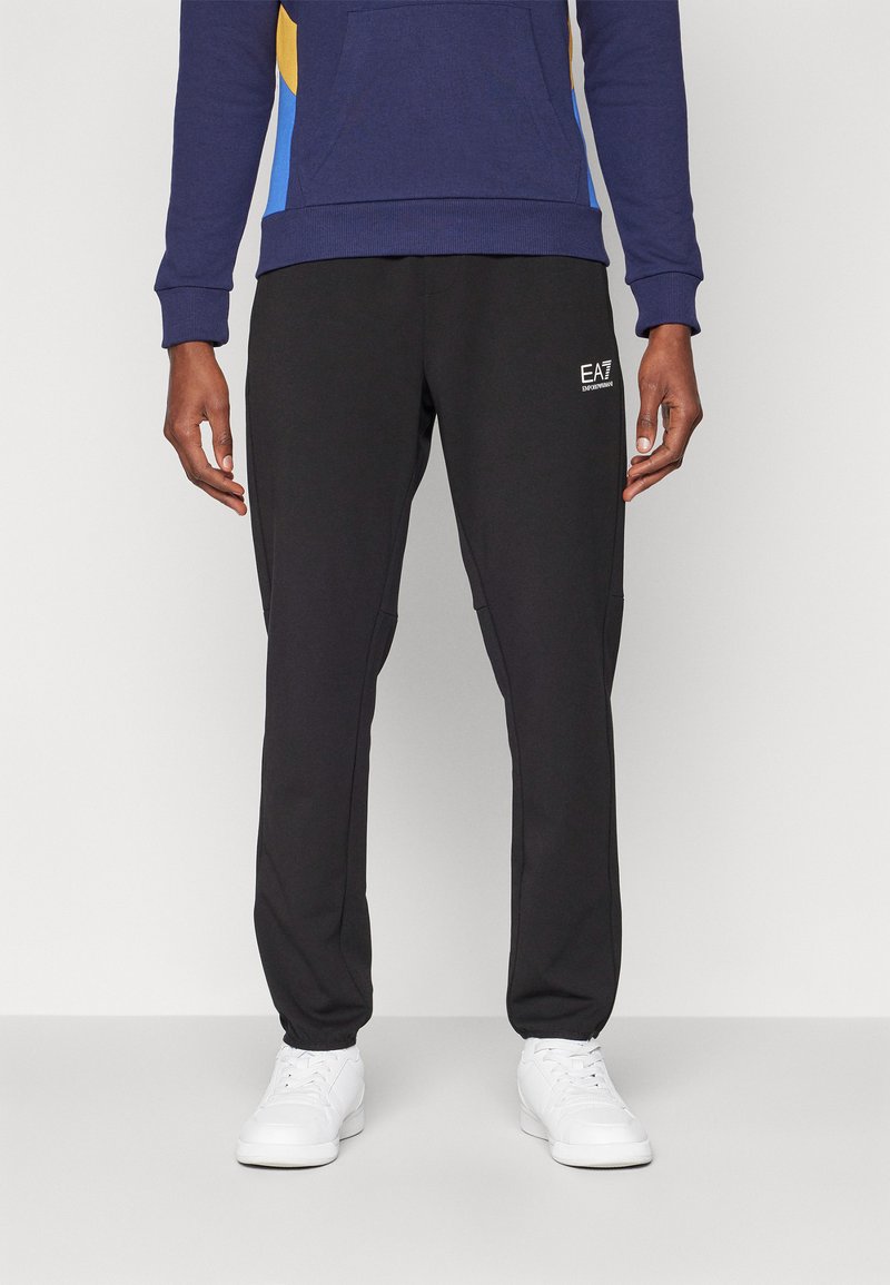 Svart joggebukse med sidelommer, innsvingt design og logodetalj. Kombinert med hvite joggesko for et komplett atletisk utseende.