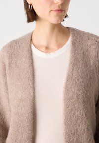 Beige, paksu kudumantel pehmea tekstuuriga, avatud ees ja langevate õlgadega. Kandmise alla on selga pandud õhuke, ribadega valge topp.