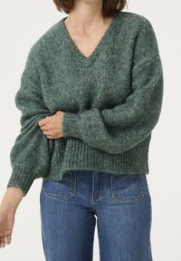 Grüner Strickpullover mit V-Ausschnitt, geripptem Saum und langen, gepufften Ärmeln. Das Material wirkt weich und strukturiert, mit einem gemischten Garn.