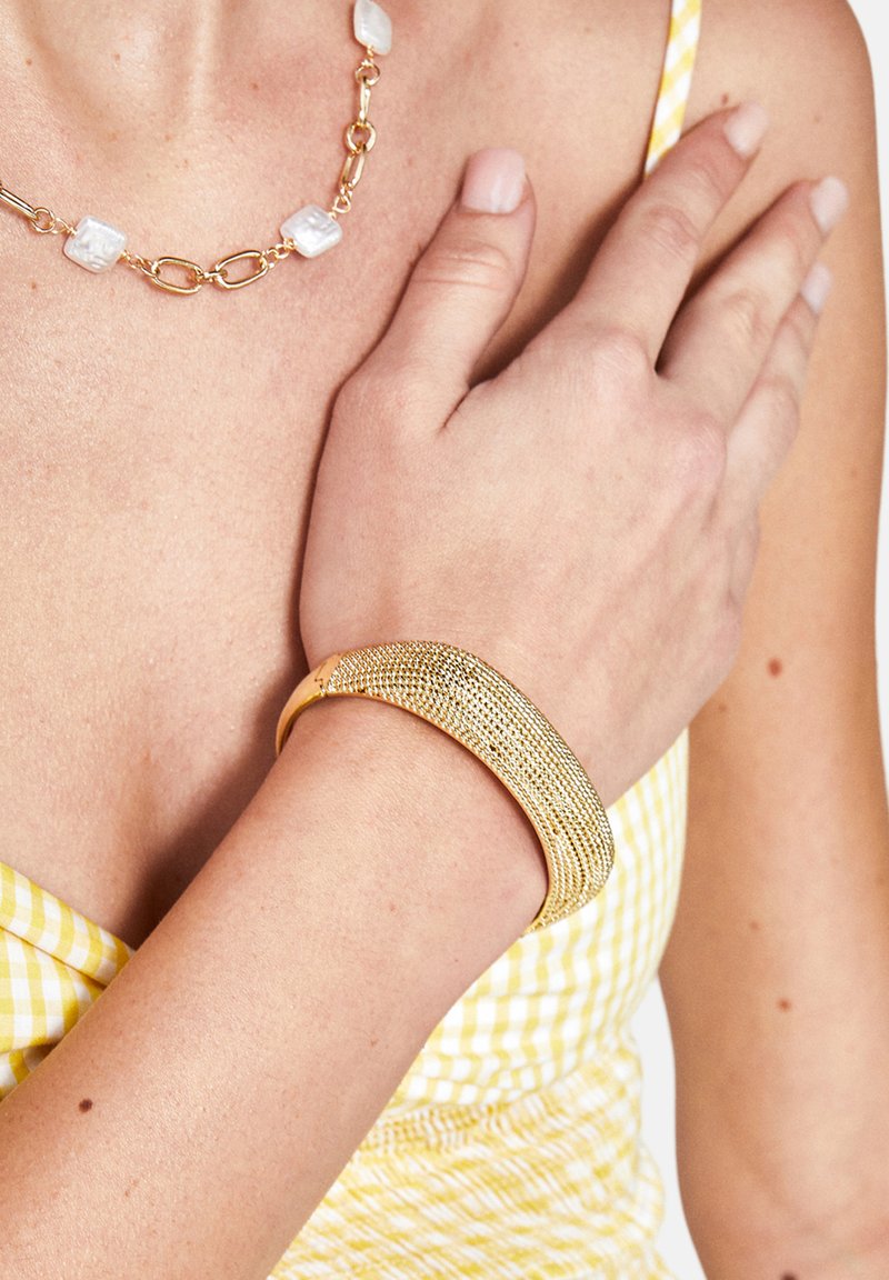 Bracciale in metallo dorato con un design testurizzato, caratterizzato da una forma allungata e curvata. Indossato su un polso contro uno sfondo giallo rigato.