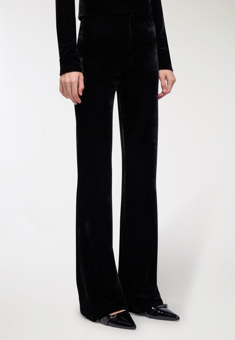 Czarne welurowe spodnie dzwony charakteryzują się gładką teksturą i wysokim stanem. Projekt ma elegancką sylwetkę i jest zestawiony z butami na spiczastym nosku.
