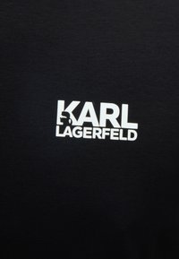 Έντονο λευκό κείμενο λογότυπου "KARL LAGERFELD" σε μαύρο υφασμάτινο φόντο, με στυλιζαρισμένη σιλουέτα ενσωματωμένη στο γράμμα "K".