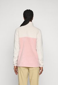 Persona con capelli intrecciati che indossa una giacca in pile color rosa pallido e bianco e pantaloni beige, in piedi con le spalle rivolte verso la telecamera su uno sfondo semplice.