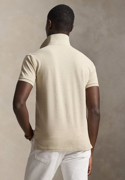 Moški, ki nosi bež polo majico s kratkimi rokavi in dvignjenim ovratnikom ter bele hlače, stoji z eno roko v žepu, z zadnjim delom telesa proti kameri.