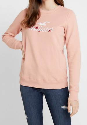 Žena nosící světle růžovou mikinu Hollister s květinovou výšivkou a tmavě modré roztrhané skinny džíny, stojící s rukou v bok.