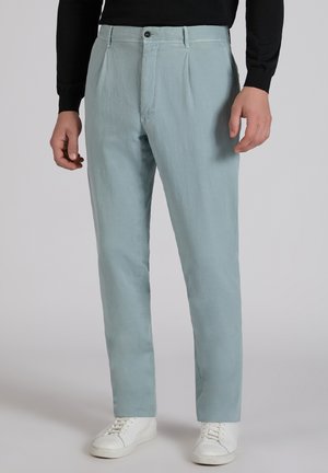 Mężczyzna w jasnoniebieskich plisowanych spodniach na guziki i suwak, w połączeniu z białymi trampkami i czarną bluzką z długim rękawem.