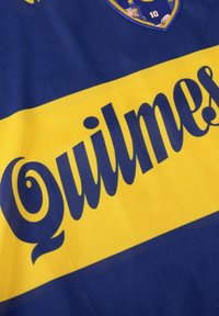 Blauw en geel stoffen sportshirt met de tekst "Quilmes" in vetgedrukte, gestileerde letters, met een schild en het nummer "10" boven de tekst.