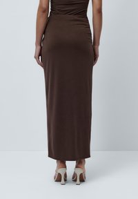 Brun maxi kjole med fitted bodice, samlet detalj bak, og et glatt, elastisk stoff. Kombinert med lyse høye hæler.