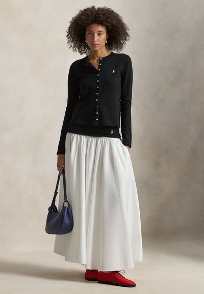 Μαύρη μακρυμάνικη ζακέτα με κουμπιά, φορεμένη πάνω από μαύρο τοπ. Λευκή φούστα που ρέει, κόκκινα παπούτσια και μπλε τσάντα συμπληρώνουν το σύνολο.