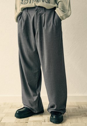 Pantaloni larghi grigi a quadri abbinati a scarpe nere chunky e un maglione beige lavorato a maglia con testo, mostrati dalla vita ai piedi contro una parete semplice.