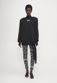 Sudadera negra de gran tamaño con hombreras, que presenta un logo. Combinada con leggings de patrón en blanco y negro. Sandalias planas.