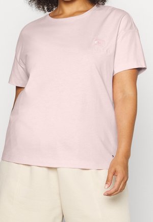 Vrouw draagt een lichtroze Tommy Hilfiger T-shirt en crèmekleurige korte broek, beide afgeknipt bij de hals en halverwege de dij, staand met één arm ontspannen langs haar lichaam.