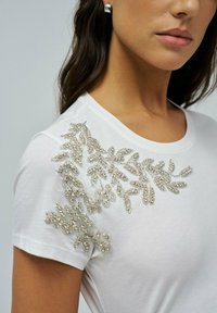 Camiseta blanca con intrincados adornos de hojas de cuentas plateadas en el hombro izquierdo, cuello redondo y mangas cortas.