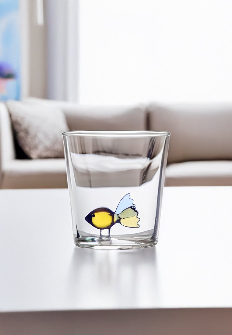Przezroczysty szklany kubek z kolorowym wzorem ryby, która ma żółte ciało, niebieskie płetwy i zielony ogon. Gładka faktura, okrągły kształt.