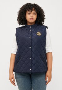 Una giovane donna con capelli ricci indossa un gilet trapuntato blu navy con un logo, abbinato a una camicia bianca e a jeans, su uno sfondo semplice.