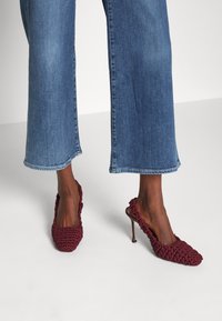Pies que usan zapatos de tacón alto burgundy trenzados tipo slingback, combinados con unos jeans de mezclilla azul cortos sobre un fondo blanco.