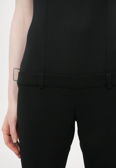 Top noir ajusté en tissu lisse, avec un design structuré, une taille plate avec des passants sur les côtés, et une silhouette slim.