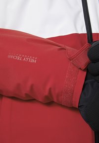 Manche de veste imperméable rouge avec ajustement velcro, arborant un logo blanc. Tissu texturé noir visible en dessous.
