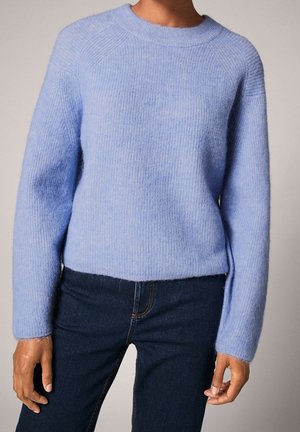 Personne porte un pull doux bleu clair à manches longues associé à un jean bleu foncé, debout devant un fond neutre.