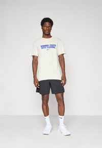 Lys beige t-shirt med blå tekst "VINDEN STARTER MED TRÆNING" og Nike-logo, parret med sorte atletiske shorts og hvide sneakers.