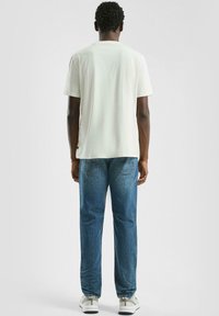 Man die met zijn rug naar ons toe staat, gekleed in een eenvoudig wit T-shirt, blauwe jeans en witte sneakers, tegen een effen lichte achtergrond.