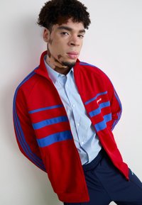 Giacca rossa con zip e strisce blu sulle maniche, indossata sopra una camicia button-up azzurra chiara. Tessuto morbido, vestibilità casual e polsini a coste.