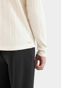 Cremfarbener, gerippter Strickpullover mit langen Ärmeln, der eine dezente Struktur und ein kleines Logo in der Nähe des Saums aufweist, kombiniert mit schwarzen, plissierten Hosen.