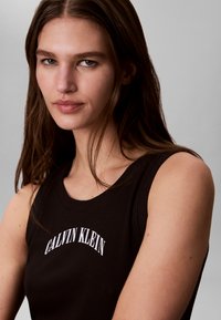 Νεαρή γυναίκα με μακριά καστανά μαλλιά, που φοράει μαύρο αμάνικο τοπ με το λογότυπο "Calvin Klein" στο στήθος, κοιτάζει ελαφρώς προς την κάμερα.