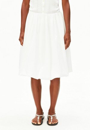 Femme portant une jupe blanche longueur genou avec taille élastique et des sandales blanches en corde, debout devant un fond blanc uni.