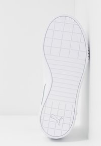 Sula av en vit Puma-sneaker med ett galler- och randmönster, placerad stående mot en vit hörnvägg.