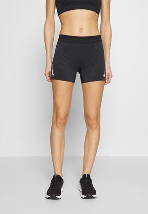 Vrouw draagt zwarte geribbelde sportbroekjes met witte logo's op beide benen en zwarte hardloopschoenen, staand tegen een witte achtergrond.