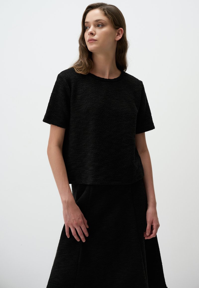 Schwarz strukturiertes Crop-Top mit kurzen Ärmeln und rundem Halsausschnitt, kombiniert mit einem passenden schwarzen Rock. Schlichte, klare Linien und minimalistisches Design.