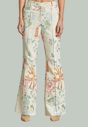 Beige Schlaghosen mit buntem Blumenstickerei, getragen mit beigen Schuhen, bis zur Taille bis zu den Füßen vor einem schlichten Hintergrund gezeigt.