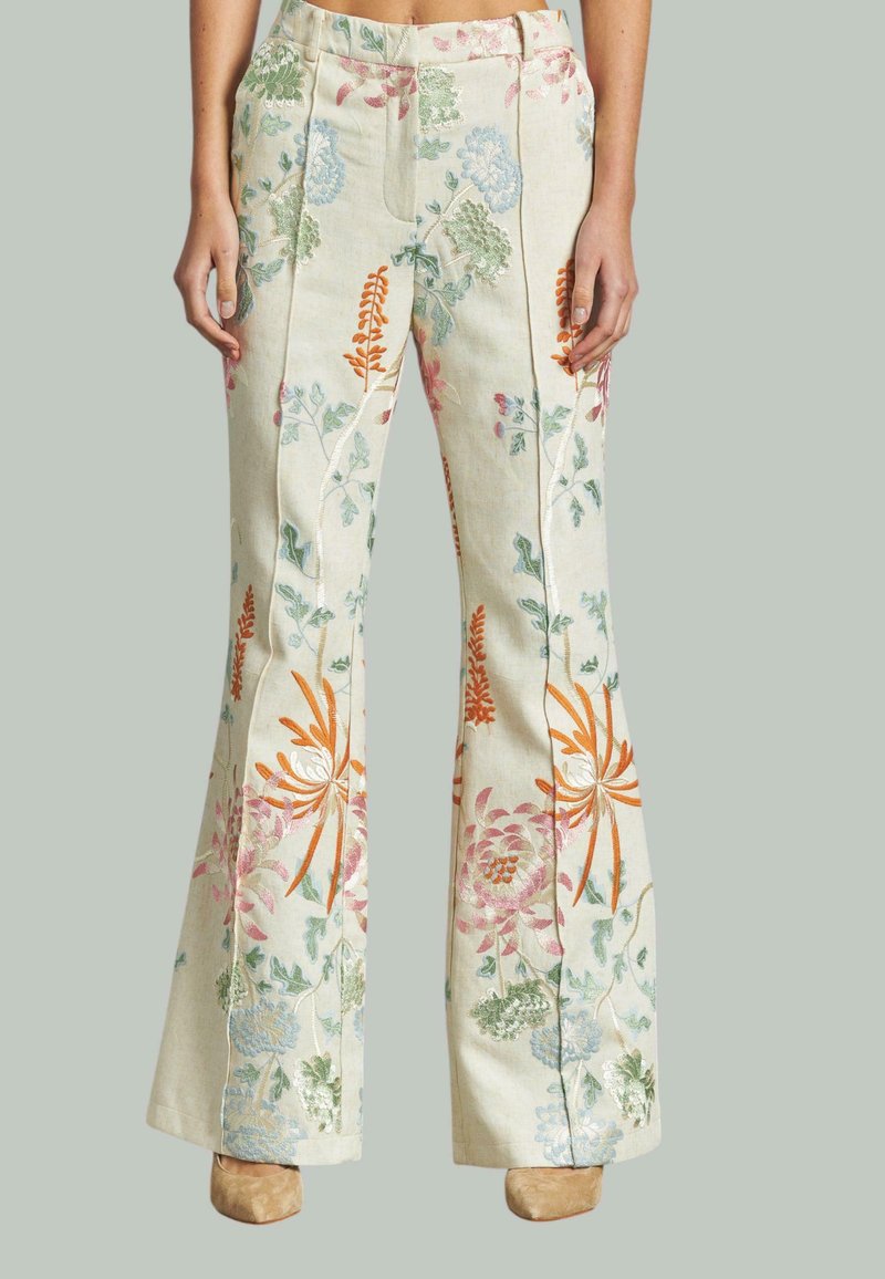 Pantaloni beige a palazzo con ricami floreali colorati, indossati con scarpe beige, mostrati dalla vita ai piedi su uno sfondo neutro.