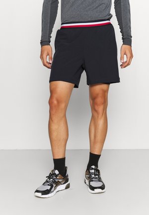 Mężczyzna ubrany w czarne sportowe spodenki z pasem w kolorach czerwonym, białym i niebieskim, szarą koszulkę z długim rękawem, czarne skarpetki i szare buty do biegania.