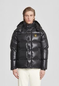 Giacca puffa nera con superficie lucida, design trapuntato, colletto alto e cappuccio attaccato. Presenta una piccola etichetta logo sul petto.