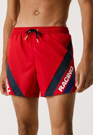 Bañador rojo con un panel lateral negro, con un diseño diagonal y la palabra "RACING" en blanco. Cintura elástica con un cordón negro.
